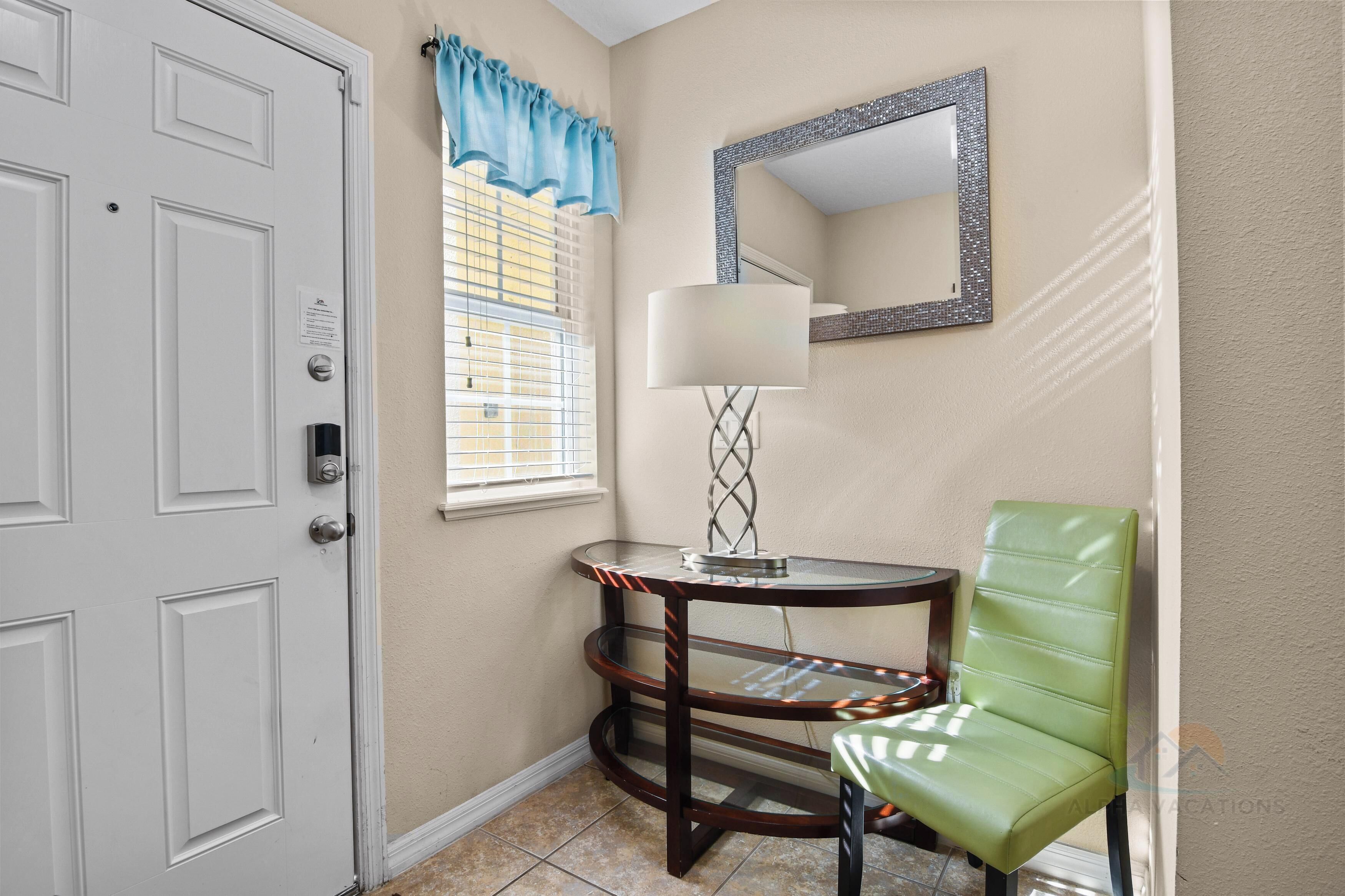 A corner room with a white door, a small table with a lamp, a green chair, and a window with blue curtains.