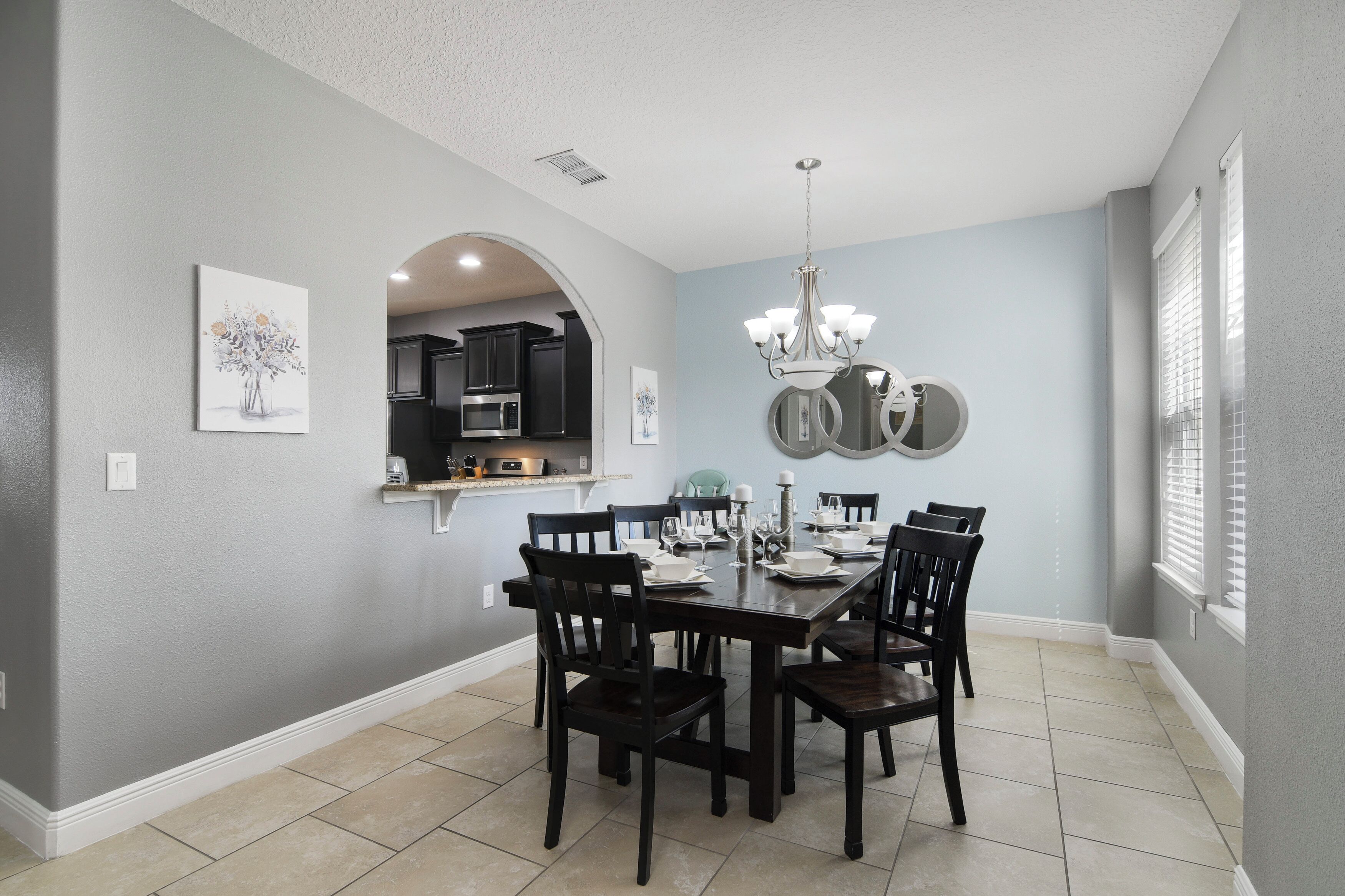 A dining room with a table set for six, a chandelier, and kitchen cabinets visible through an archway.