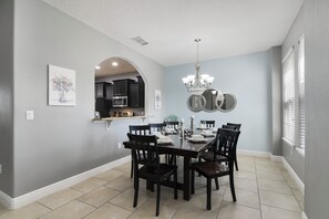 Une salle à manger avec une table dressée pour six personnes, un lustre et des meubles de cuisine visibles à travers une arche.