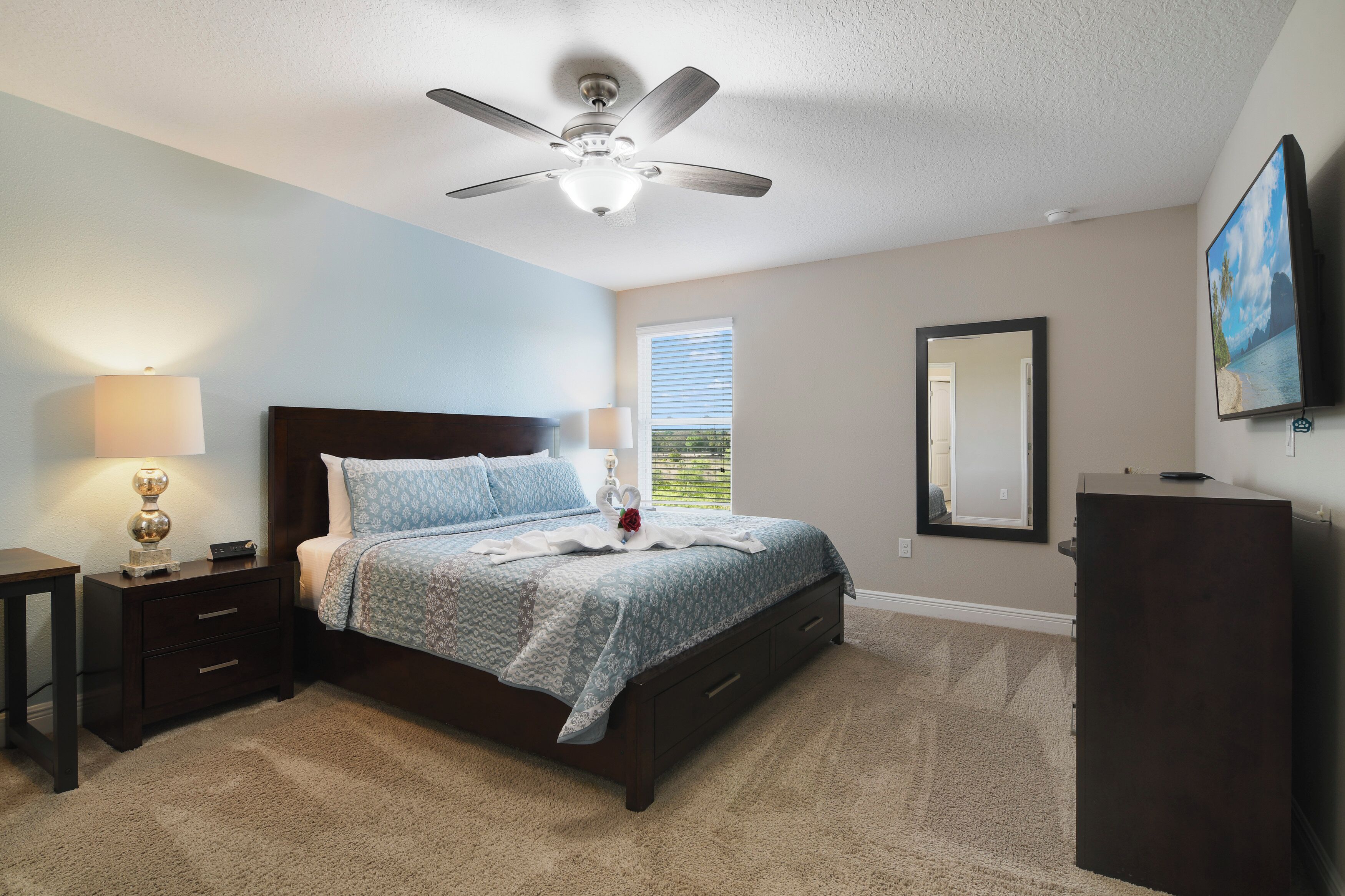 A bedroom with a bed, nightstand, lamp, ceiling fan, window, and a TV on a stand.