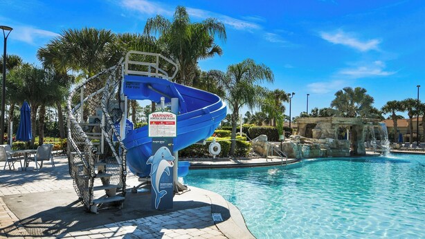 Un espace piscine doté d’un toboggan aquatique aux couleurs bleues, de palmiers et d’un ciel d’un bleu limpide.