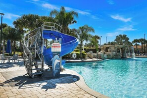 A pool area with a blue water slide, palm trees, and a clear blue sky. - Theatre & Games - Luxe Resort +10 Min Disney +pets (Kissimmee)