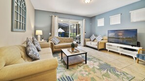 A living room with a sofa, armchair, coffee table, TV, and a view through a glass door. - Sunset Arcade - Large Pool/spa + Luxe Resort +pets (Davenport)