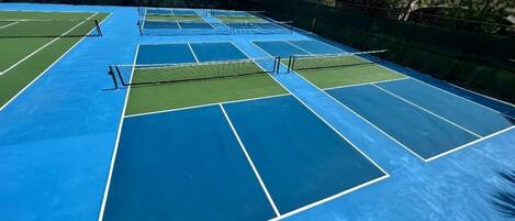 A tennis court with multiple courts, surrounded by a green fence and trees.
