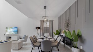 A modern dining area with a round table set for four, surrounded by grey upholstered chairs with gold legs. A large flat-screen TV is mounted on the wall to the left, and a floor lamp is positioned to the right of the table. - Champions Oasis - Resort&arcade -walk to Clubhouse (Davenport)