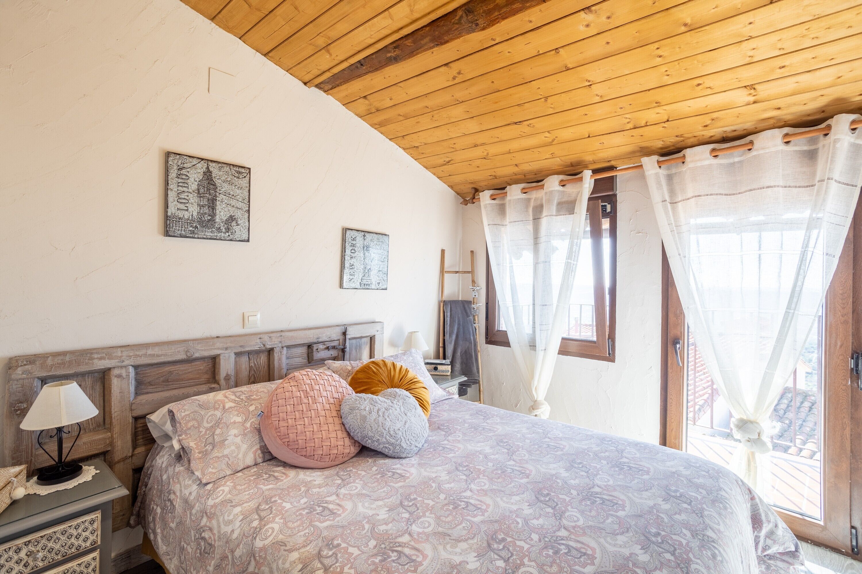 Un dormitorio con techo de madera, una cama con ropa de cama estampada, una mesita de noche con lámpara y una ventana con cortinas de gasa.