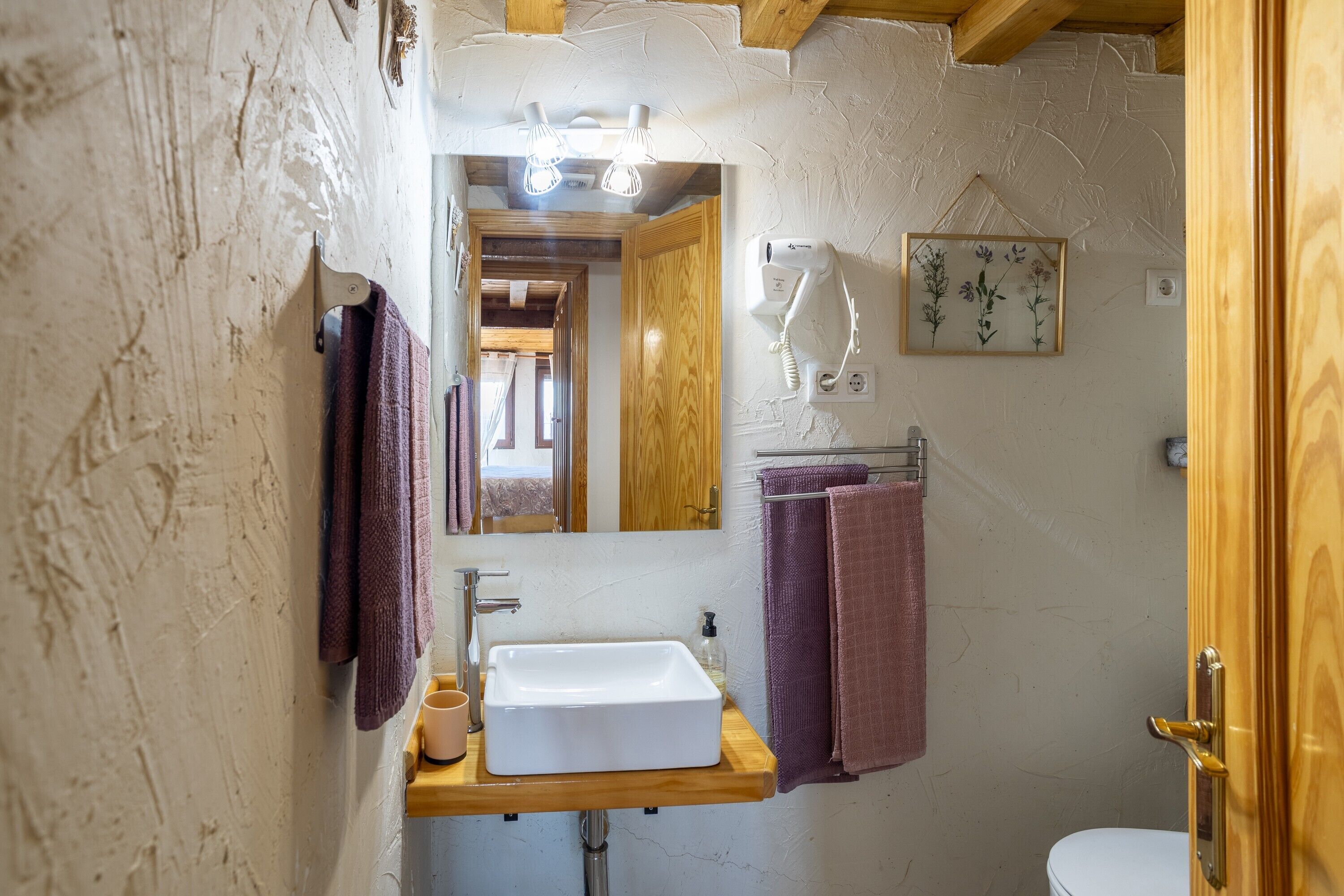 A bathroom with a wooden vanity, a white sink, a mirror, and a framed floral picture on the wall.