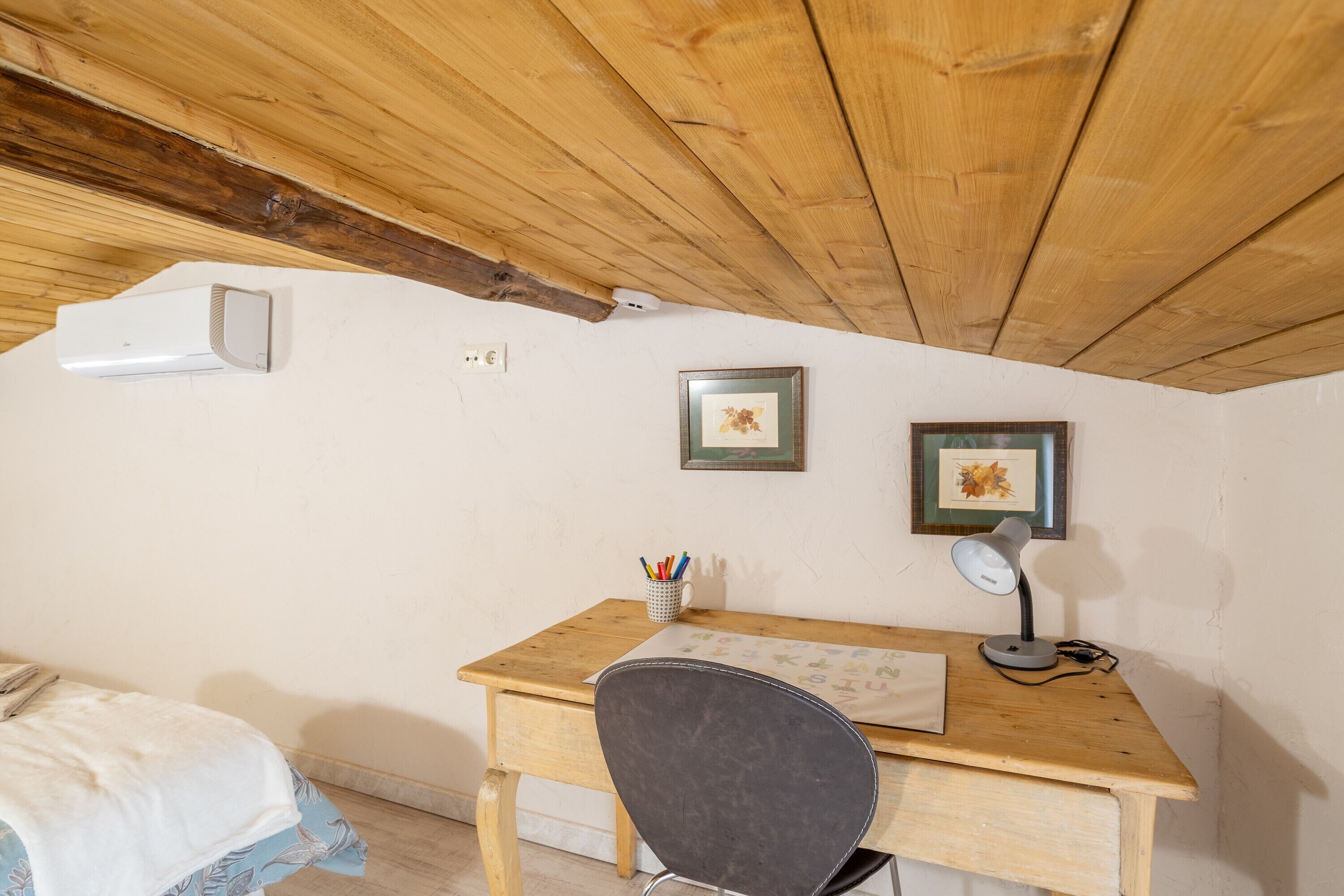 A room with a wooden desk, a chair, a bed with a white blanket, and two framed pictures on the wall.