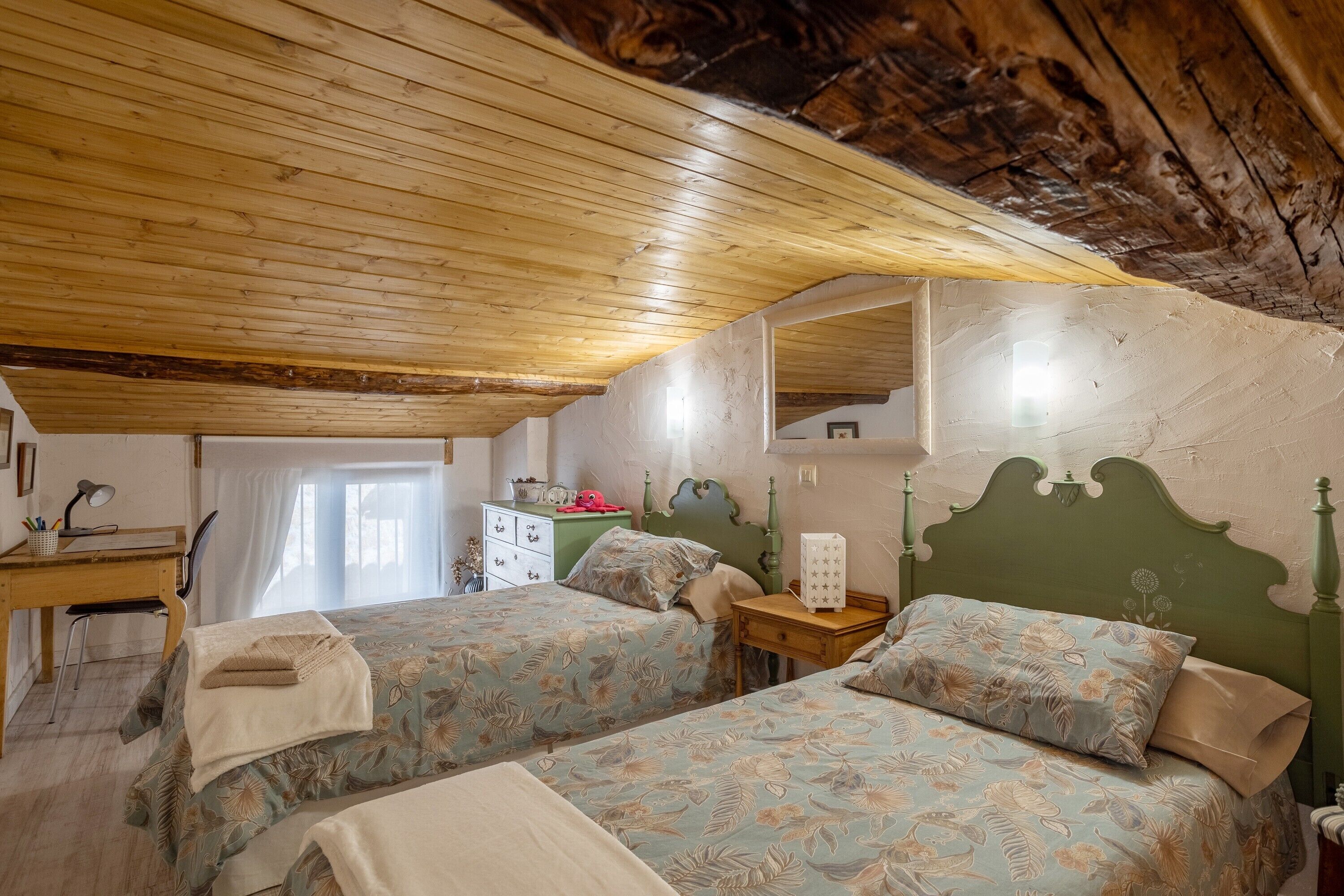 A room with two beds, a wooden ceiling, a desk, and a dresser.