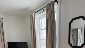 Een kleine kamer met een houten bureau, een zwarte stoel, een televisie en een raam met gordijnen.