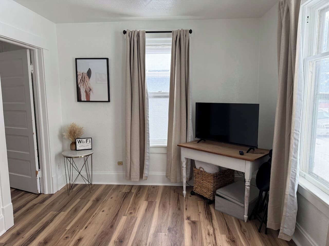 A room with a wooden floor, a TV on a wooden stand, a small table with a Wi-Fi sign, and a window with curtains.