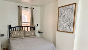Een slaapkamer met een bed, houten vloer, een raam met gordijnen, een nachtkastje en een decoratief wandobject.