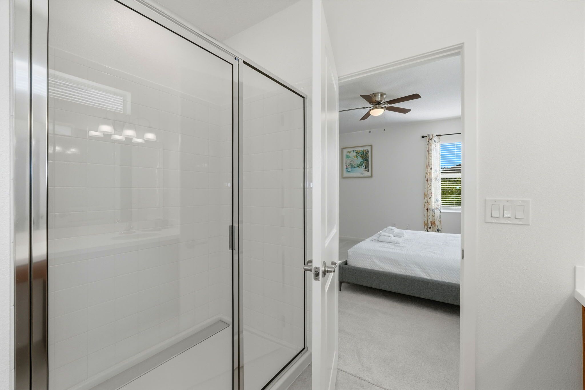 Un dormitorio con cama, ventilador de techo, ventana con cortinas y puerta que da a un baño con cabina de ducha de vidrio.