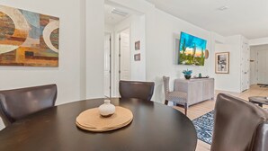 Een eetgedeelte met een ronde tafel, stoelen en een vaas. Aan de muur hangt een schilderij en er is een deur naar een andere kamer.