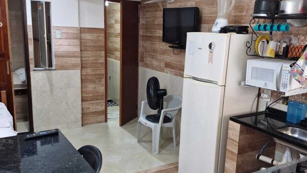 Eine kompakte Küche mit Kühlschrank, Mikrowelle und Herd. Ein Esstisch mit Stühlen und eine kleine Sitzecke mit einem Sessel sind vorhanden. Ein Fernseher ist an der Wand befestigt, und im Hintergrund ist ein Fenster zu sehen.