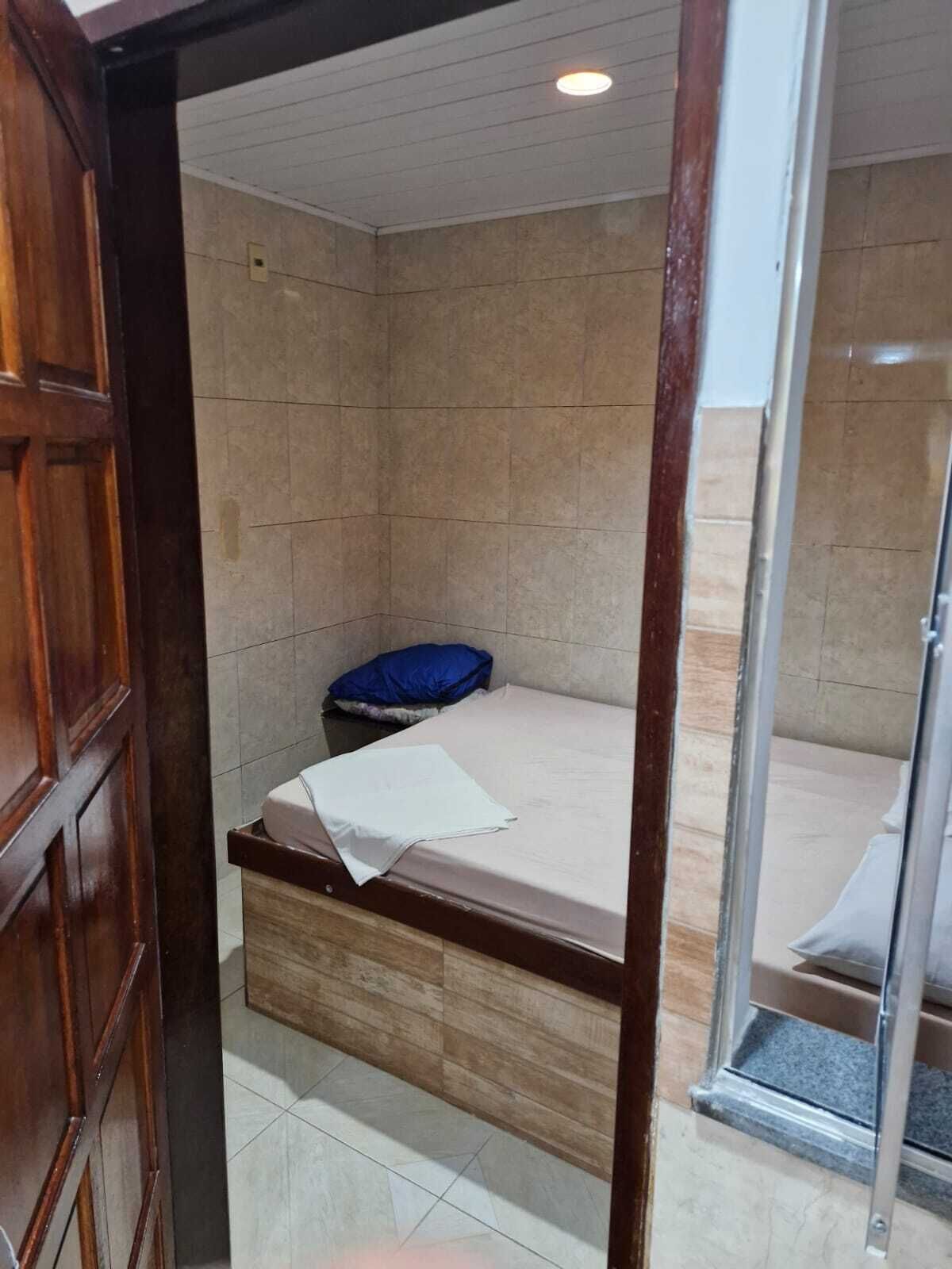 Um quarto de hotel pequeno e simples, com porta de madeira, uma cama com lençol branco e travesseiro azul, e uma mesinha.