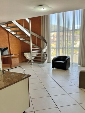 A spacious living area with a spiral staircase, a black leather chair, and a large window offering a view of the outdoors. - Apartamento 201 Saint Sebastian Flat (Jaraguá do Sul)