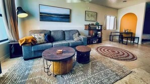 Een woonkamer met een bank, twee ronde bijzettafeltjes, een kamerkleed met patroon, een schilderij en een klein tafeltje met decoratieve objecten.