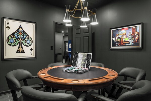 A poker table with chairs, a framed playing card, and a poker chip box in a room with dark walls.
