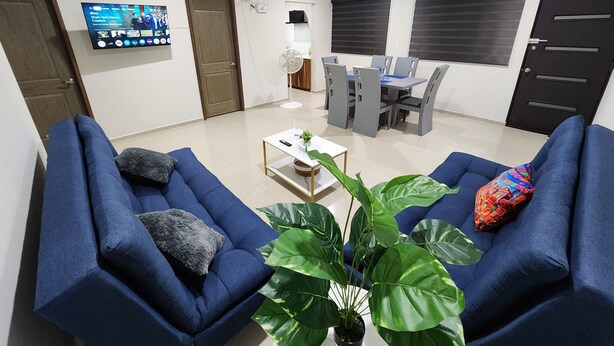 A living room with two blue sofas, a coffee table, and a large plant. There is a TV mounted on the wall and a dining area with a table and chairs in the background. - Central Apt, Ground Floor, 24h Secure Parking (Durango)