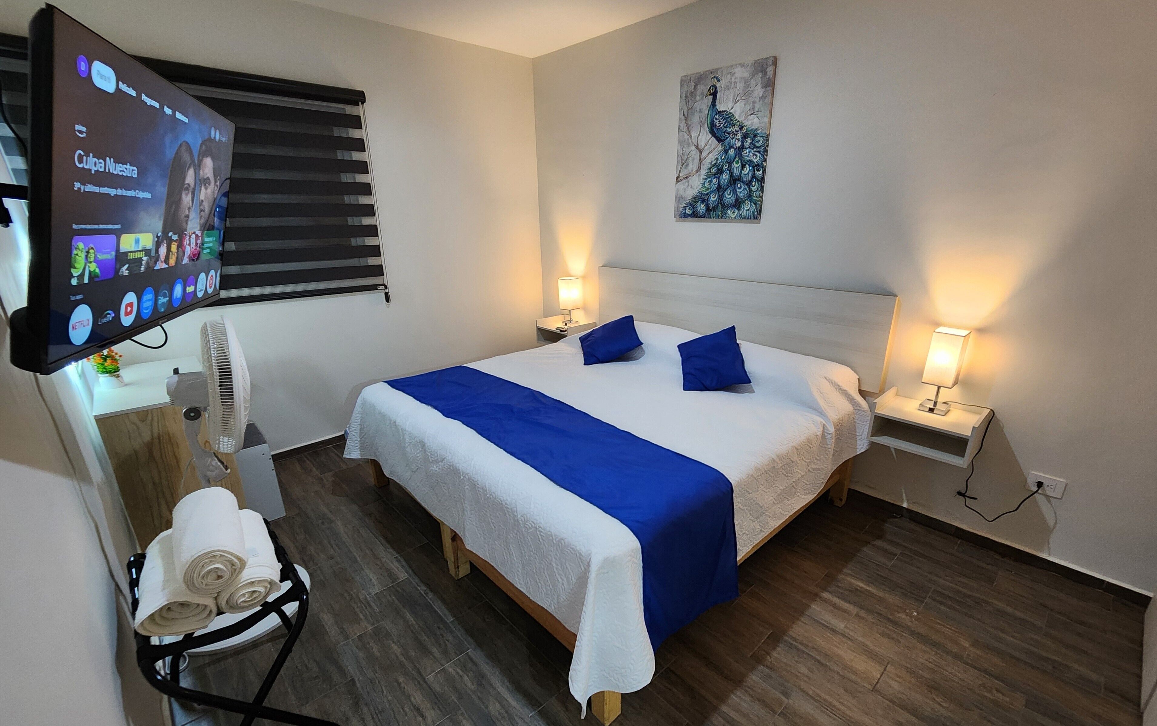 A hotel room with a bed, two bedside lamps, a wall-mounted TV, a ceiling fan, and a painting.