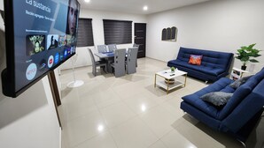 A modern living room with a flat-screen TV displaying a movie, a blue sofa, a coffee table, and a dining area with chairs and a table. - Central Apt, Ground Floor, 24h Secure Parking (Durango)