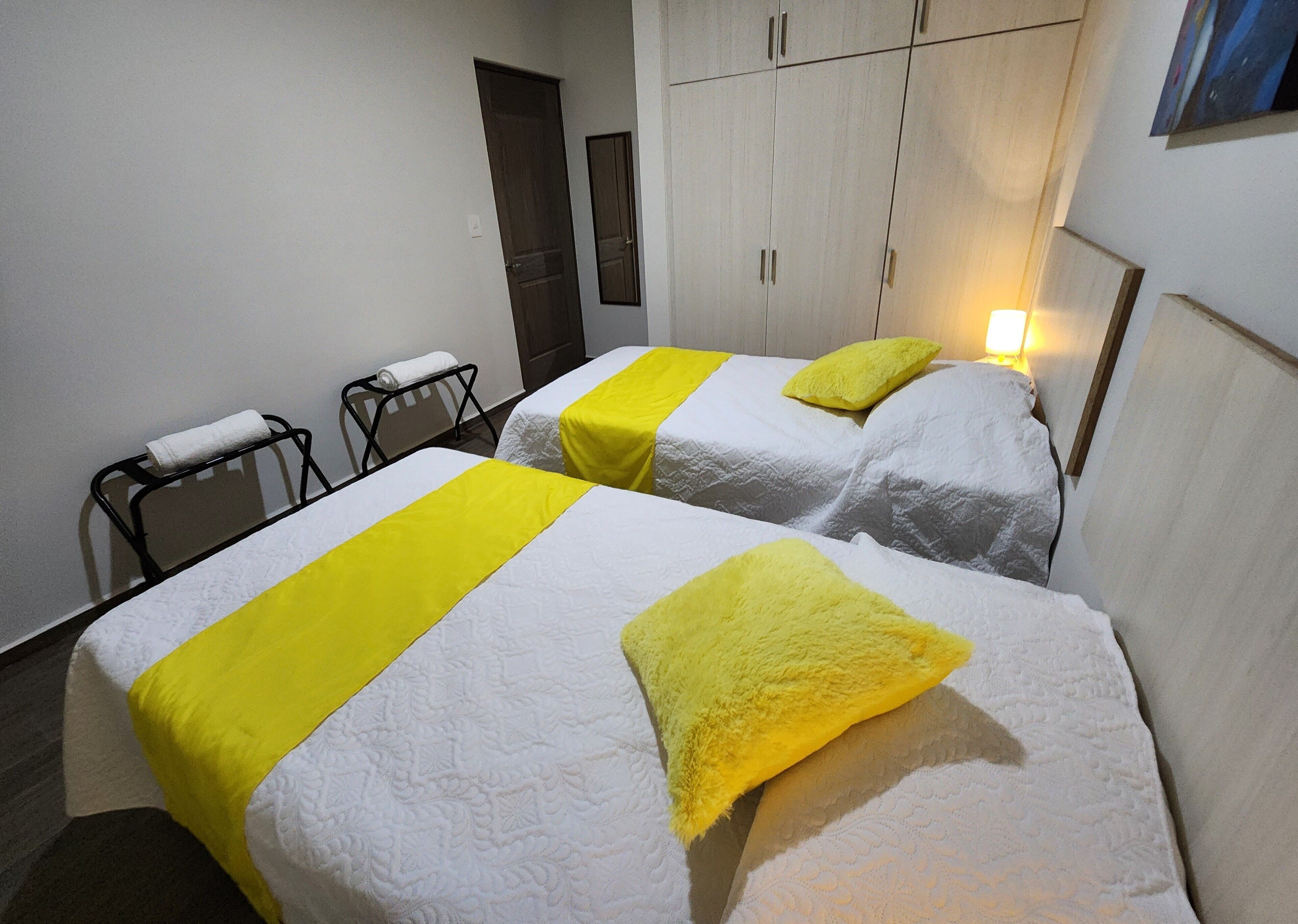 A hotel room with two beds, each with a white bedspread and a yellow accent. There is a small table with a towel on it, and a wardrobe in the background.