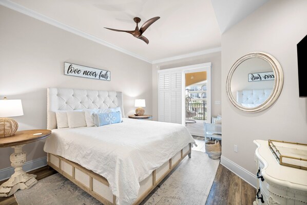 A bedroom with a tufted headboard, a ceiling fan, a bedside table with a lamp, a mirror, and a view of the outside balcony. - Paradise at 30A! 3 Bdrm plus bunks, sleeps 10-just 96 seconds to the beach! (Santa Rosa Beach)