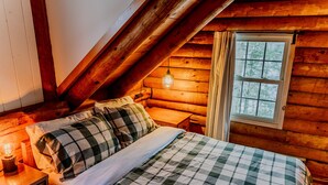 A cozy bedroom with a wooden ceiling, a bed with checkered bedding, a bedside lamp, and a window with curtains. - Rustic Log Cabin w/ Modern Updates - Close to Stowe - Steps from LV Rail Trail (Hyde Park)