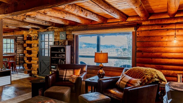 A cozy log cabin interior with a fireplace, leather furniture, and a large window offering a view of the outdoors. - Rustic Log Cabin w/ Modern Updates - Close to Stowe - Steps from LV Rail Trail (Hyde Park)
