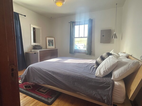 Um quarto com cama, penteadeira, espelho, janela com cortinas e luminária no teto.