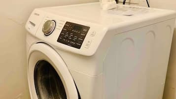 Una lavadora de carga frontal con panel de control, un cable de alimentación y un estante pequeño con una caja de pañuelos.