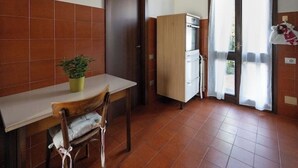 Een kleine keuken met een houten tafel, een stoel, een bloempot, een koelkast en een kast.