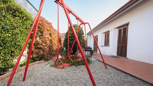 A red swing set in a courtyard with gravel ground, a white building with wooden shutters, and a hedge. - At Licia's house (Cuggiono)