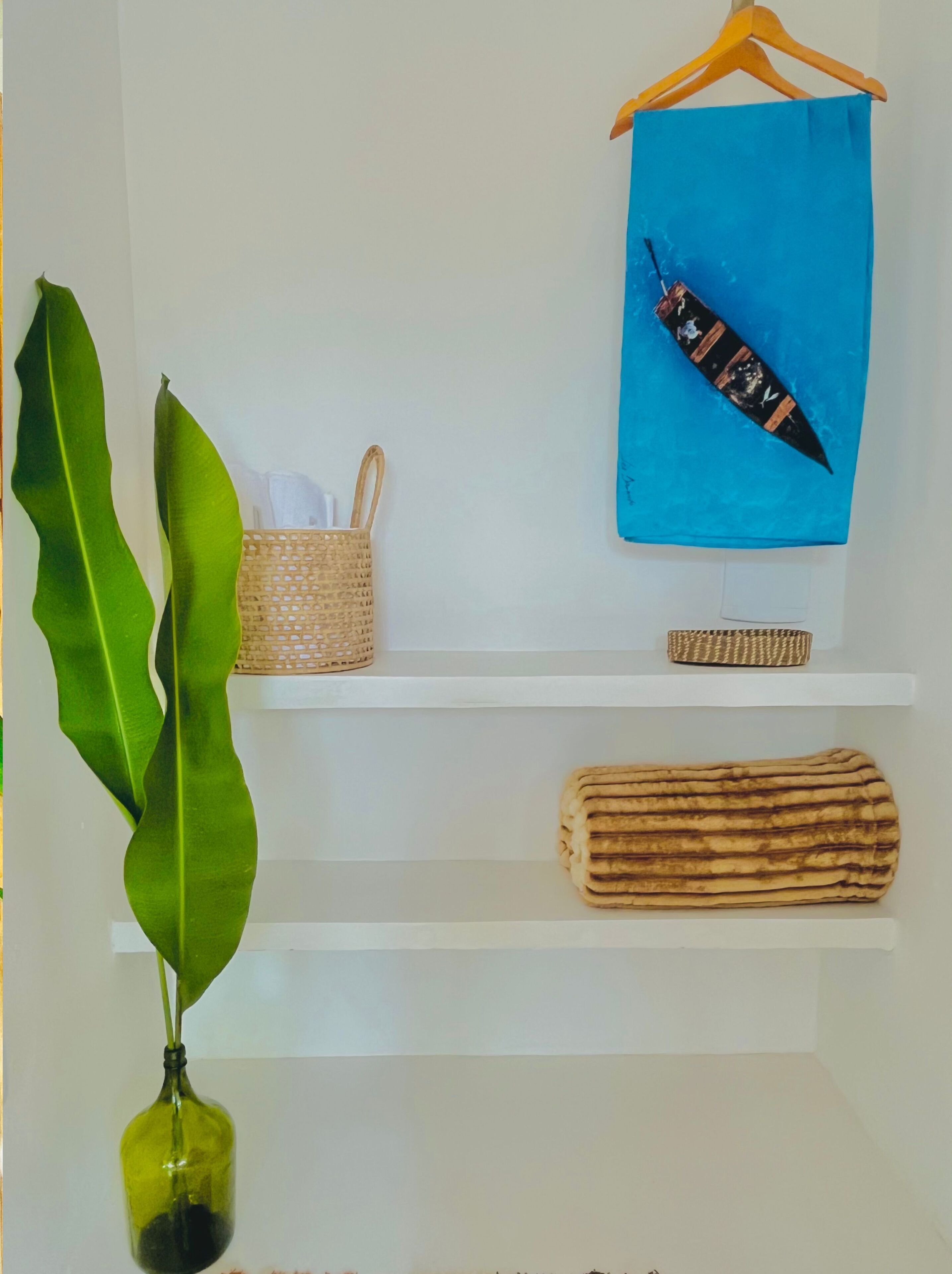 Una estantería con una cesta trenzada, un paño azul con diseño de pájaro, un objeto enrollado y una planta verde en un florero de vidrio.