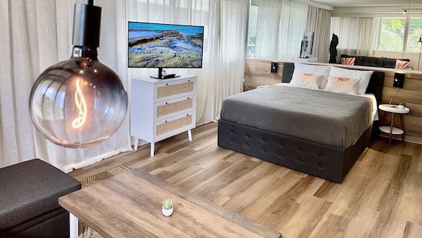 Ein modernes Hotelzimmer mit einem großen Bett, einem Fernseher auf einer weißen Kommode, einem kleinen Tisch mit einer Pflanze und einem großen Fenster mit Vorhängen.
