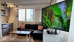 Ein modernes Wohnzimmer mit einer Couch, einem Couchtisch und einem Flachbildfernseher. Es gibt einen Küchenbereich mit einer Mikrowelle und Schränken.