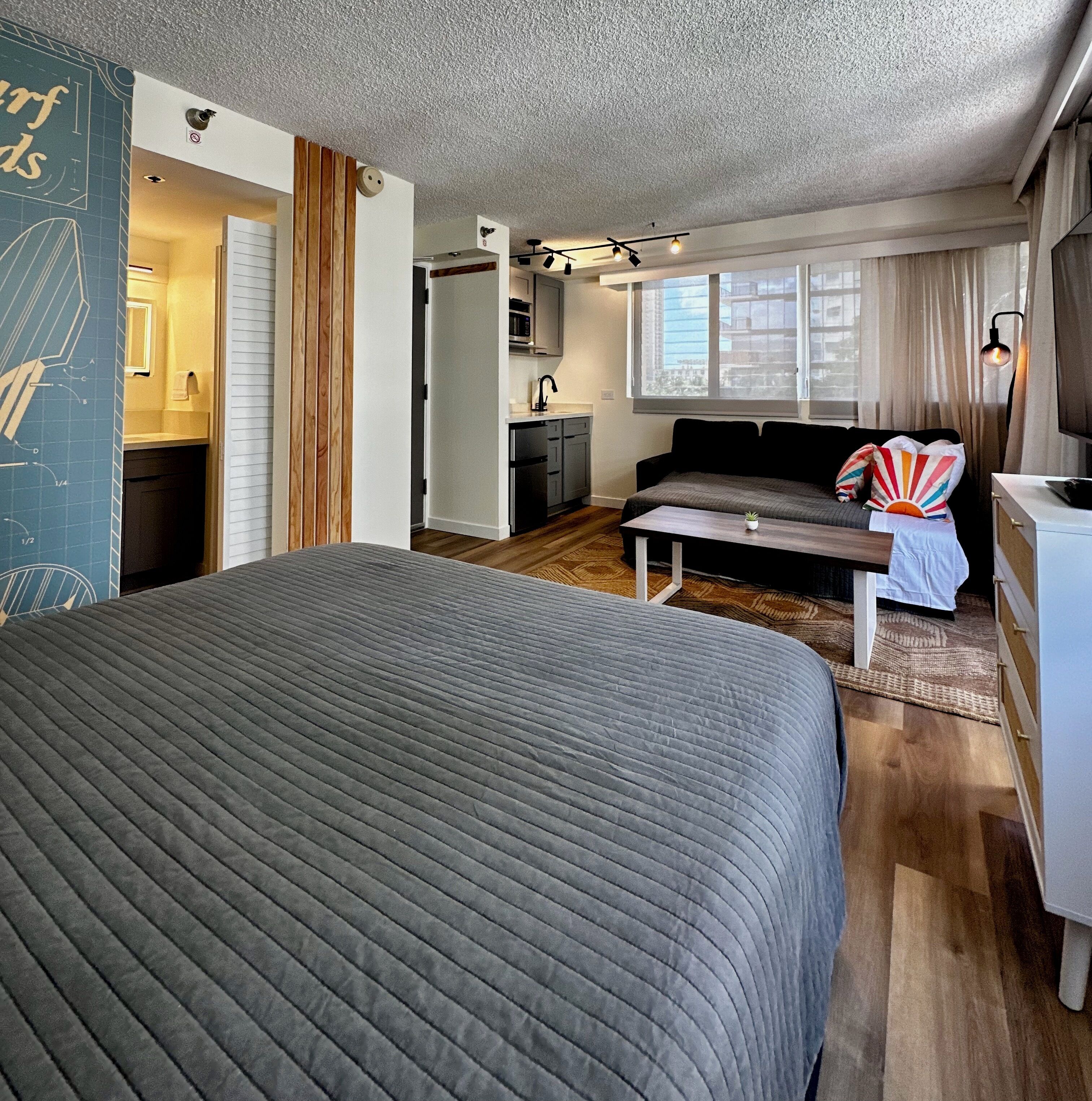 Ein modernes Schlafzimmer mit einem großen Bett, einem kleinen Tisch, einer Couch und einer Küchenzeile im Hintergrund.