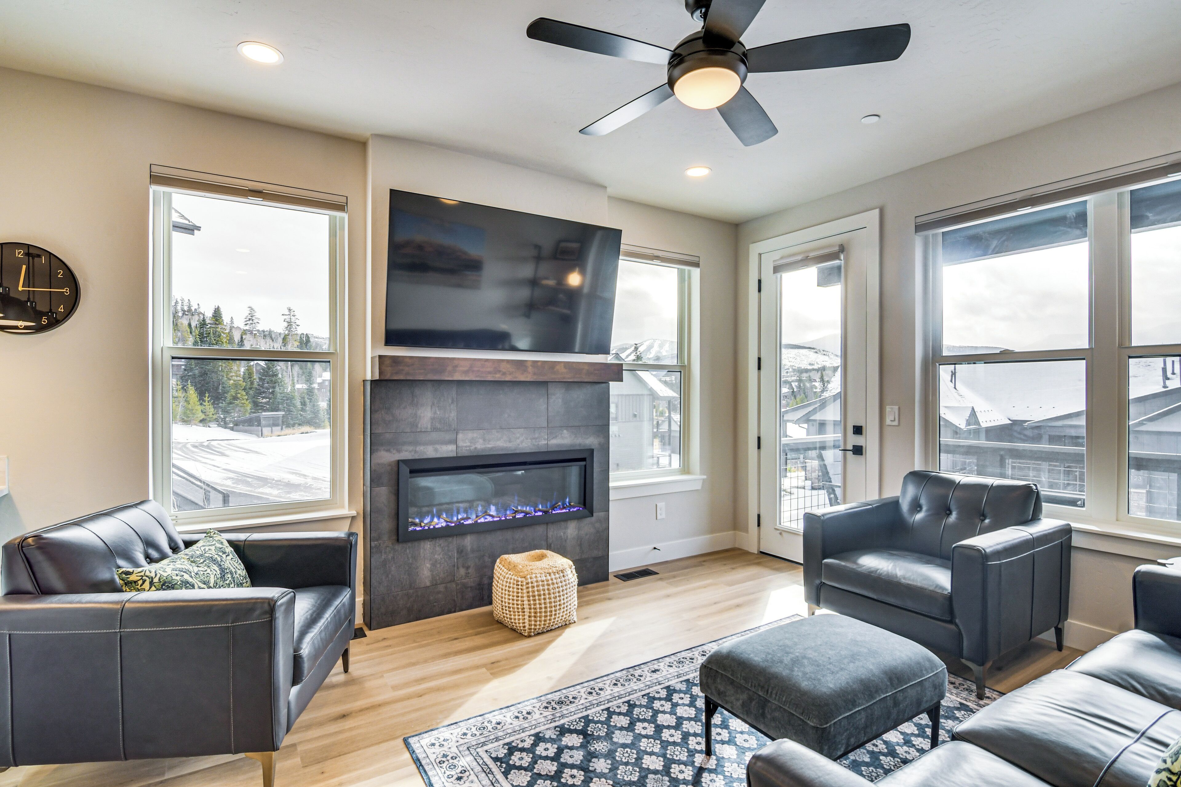 A modern living room with a fireplace, leather sofas, a ceiling fan, and large windows offering a view of the city.