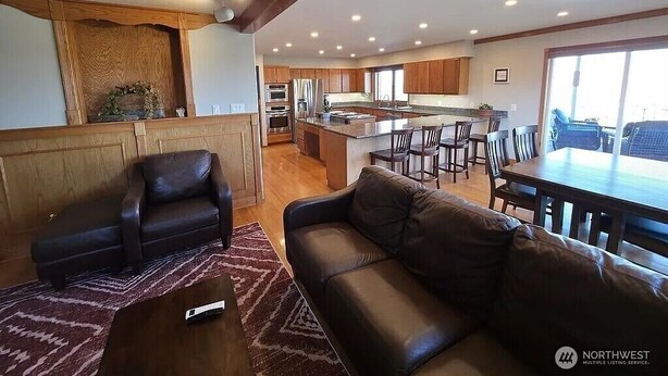 Amplia sala de estar con sofá de cuero, una mesita de centro y una mesa de comedor con sillas. Se ve una cocina con gabinetes de madera y un ventanal grande al fondo.