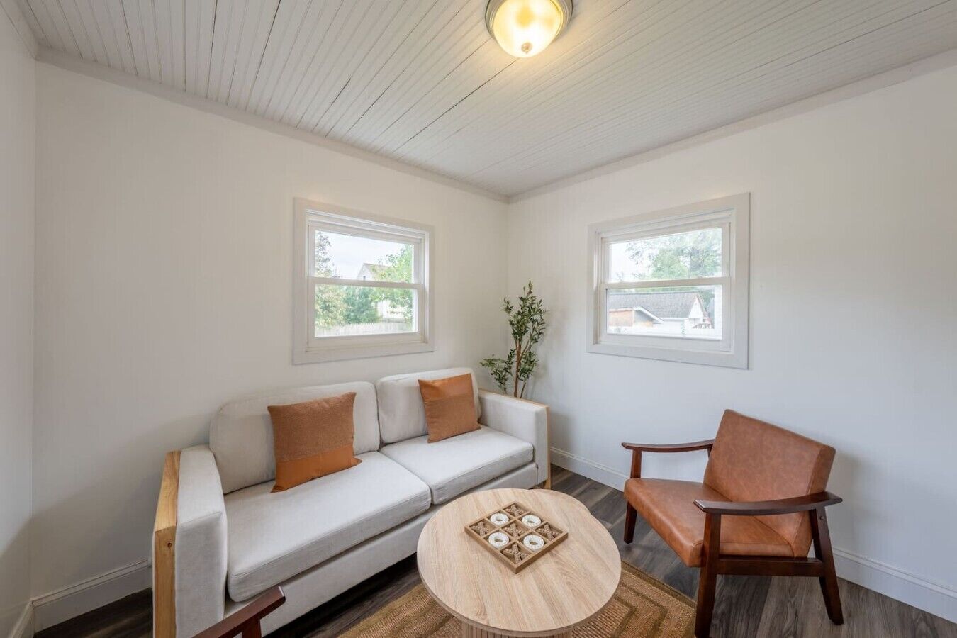 A room with a white sofa, two wooden chairs, a round wooden coffee table, and two windows with white frames.