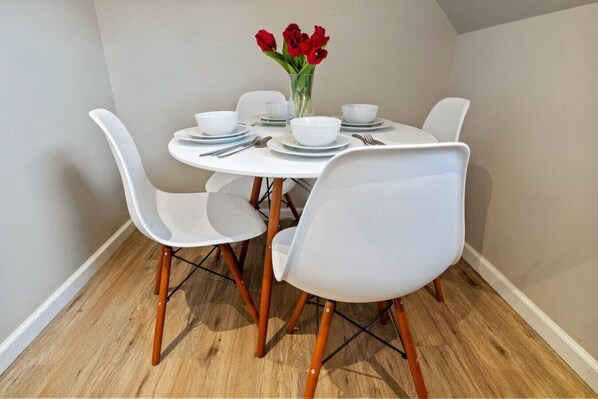 A dining area with a round table, white chairs, and a vase with red flowers.