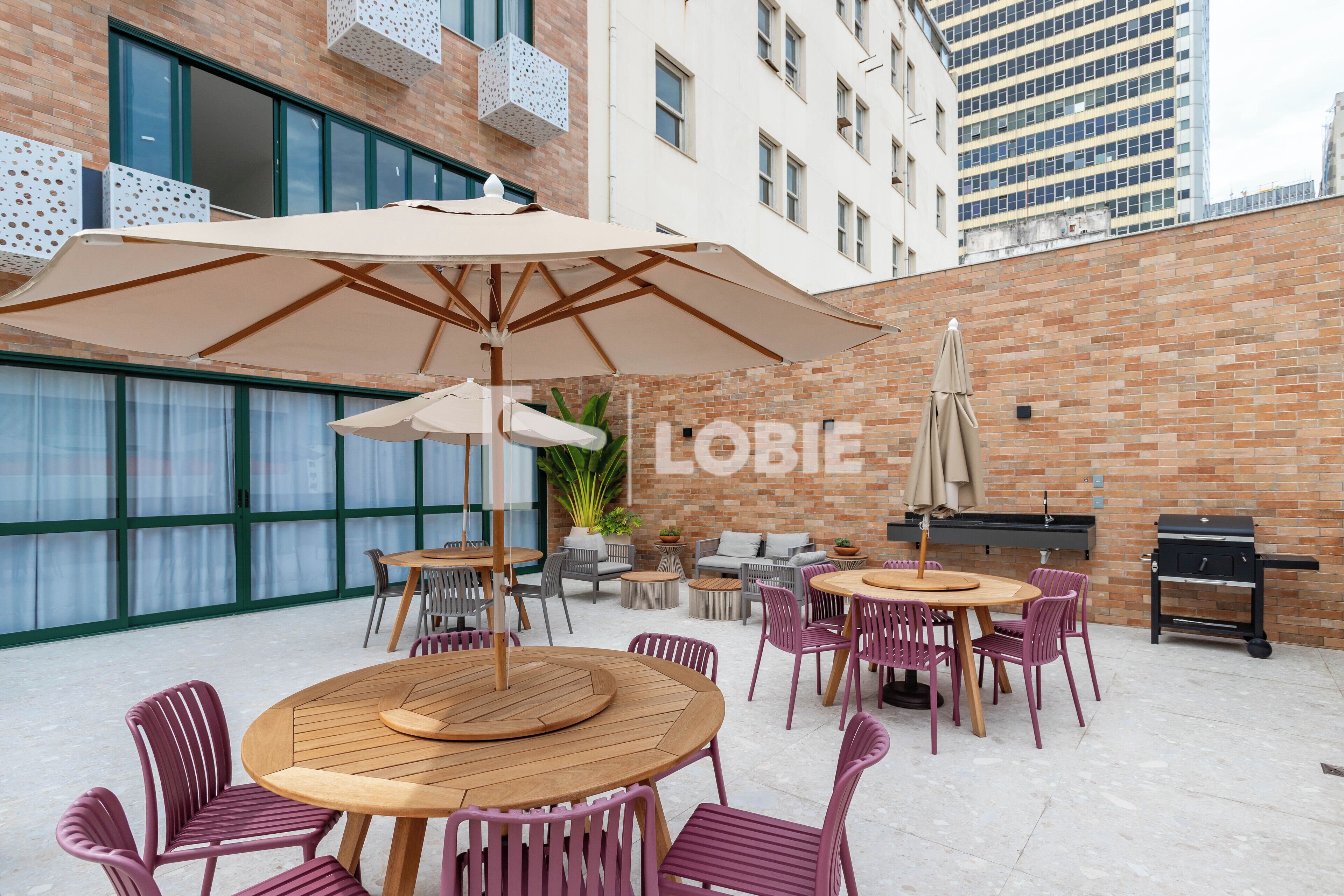 An outdoor patio with round wooden tables and purple chairs, a large umbrella, and a brick wall.