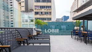 Un área de estar al aire libre con mobiliario moderno, una pared verde con la palabra "LOBIE" y un paisaje urbano al fondo.
