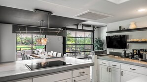 A modern kitchen with a large island, white cabinetry, a flat-screen TV, and a view of a patio with outdoor seating.