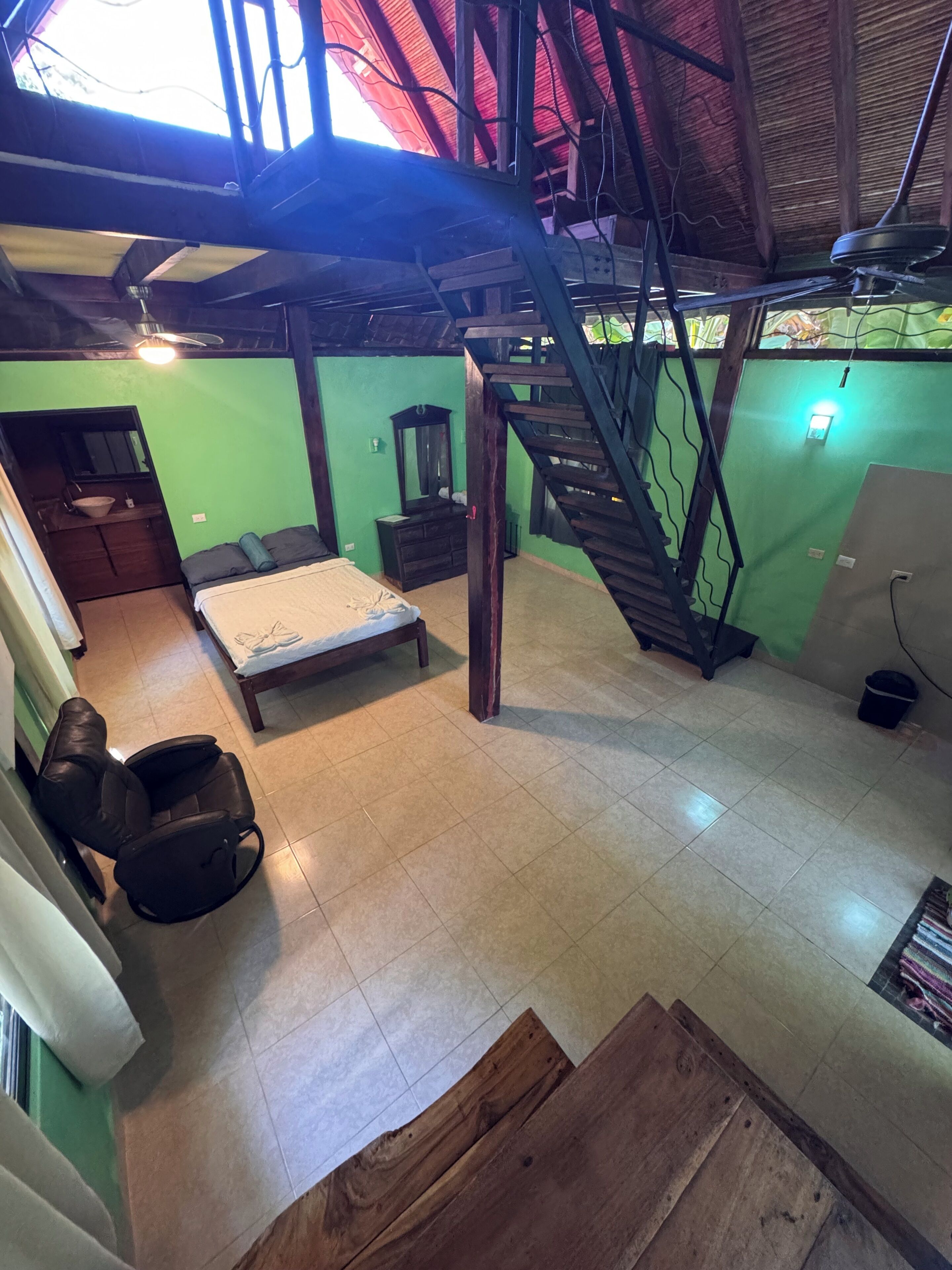 Una habitación con cama, una silla de madera, una escalera y un pequeño baño.