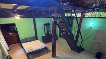 Una habitación con cama, una silla de madera, una escalera y un pequeño baño.
