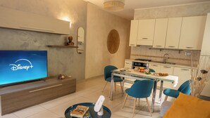 A modern living room with a flat-screen TV displaying the Disney+ logo, a dining area with a glass table and blue chairs, and a kitchen with white cabinets and a tiled backsplash. - NEW Charming 2-Bedroom Apartment in Private Roman Villa Oasis (Lazio)