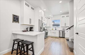 A modern kitchen with white cabinets, a stainless steel refrigerator, and a built-in oven. There are two black stools at the counter. - Lake view Townhouse | Private Hot Tub (Eden)
