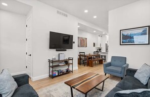 A modern living room with a flat-screen TV, a coffee table, and two sofas. There is a dining area with a wooden table and chairs. - Lake view Townhouse | Private Hot Tub (Eden)